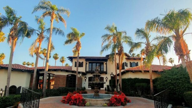 A courtyard with a fountain, palm trees, and a building with a tiled roof and stucco walls.