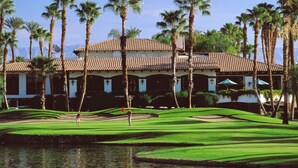 A golf course with a water hazard, palm trees, and a building with a tiled roof.