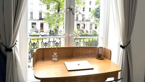 A room with a wooden table, a laptop, and a glass of water. There is a window with curtains, and a balcony railing with a view of buildings and trees.