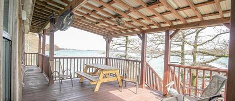 A covered deck with a wooden table and chairs, overlooking a body of water.