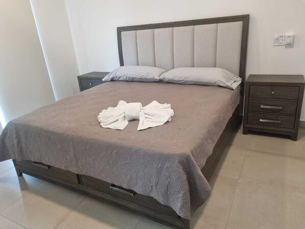 A neatly made bed with a grey bedspread and two pillows, a wooden nightstand with a drawer, and a light switch on the wall.