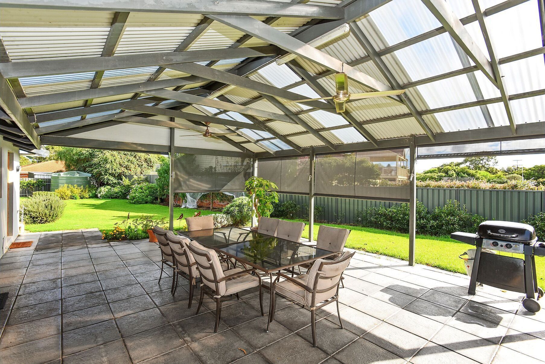 A covered patio with a glass table and chairs, a grill, and a view of a green garden.
