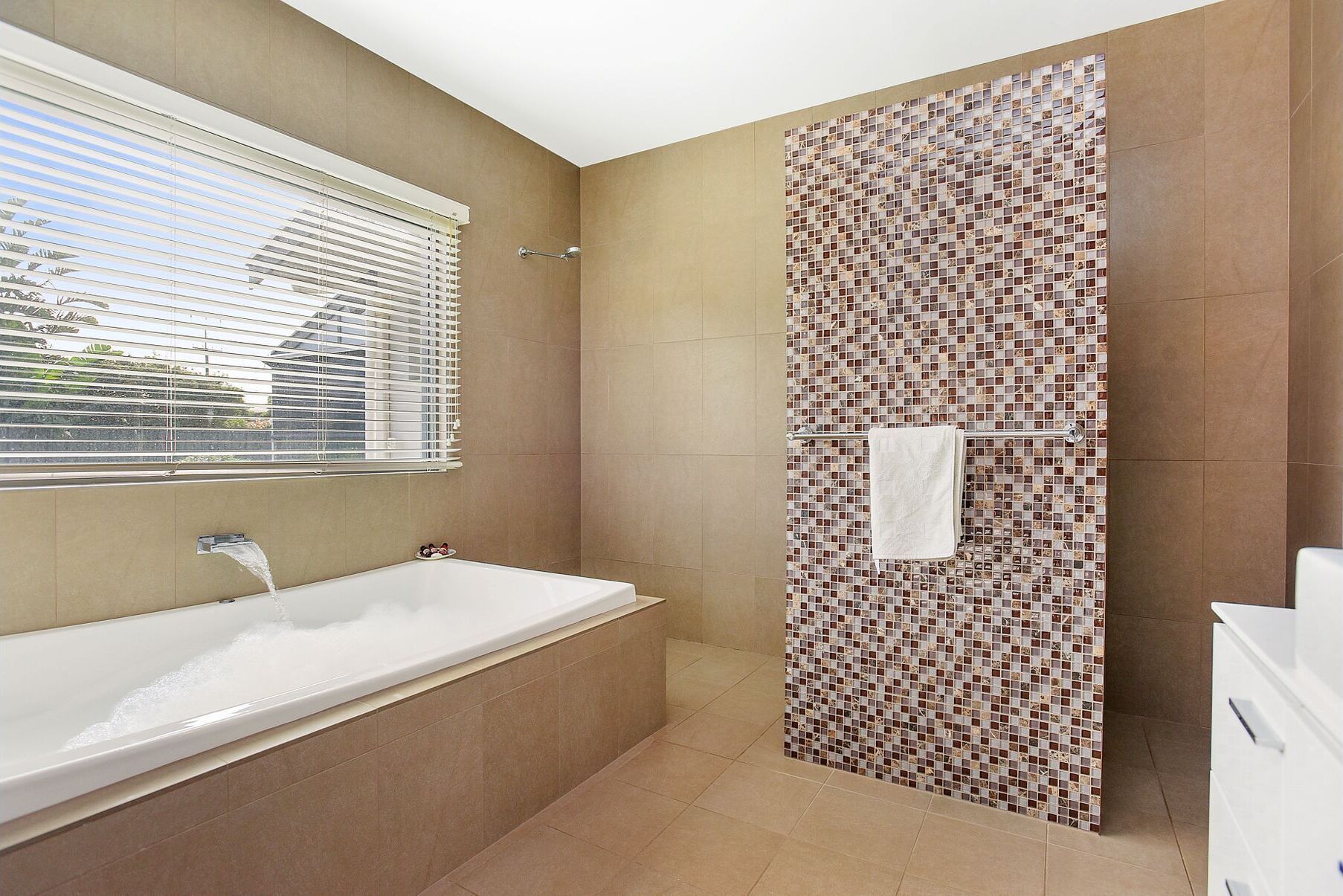 Un baño con bañera, zona de ducha con mosaicos y una ventana con persianas.