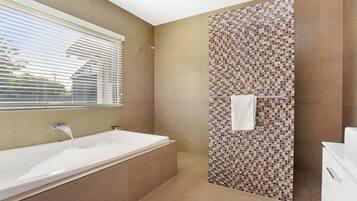 Un baño con bañera, zona de ducha con mosaicos y una ventana con persianas.