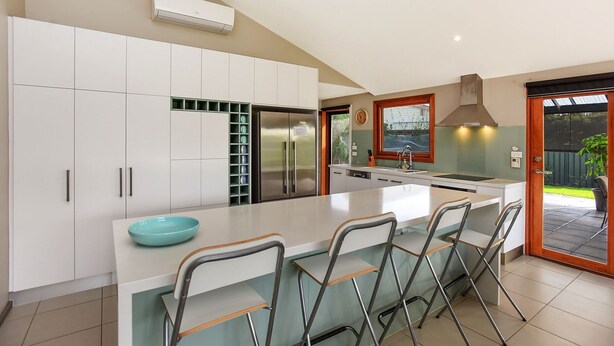 A modern kitchen with white cabinets, a stainless steel refrigerator, a large island with bar stools, and a window overlooking a garden. - Riverport Retreat - Pet Friendly, close to the river, free Wifi + linen included (Goolwa South)