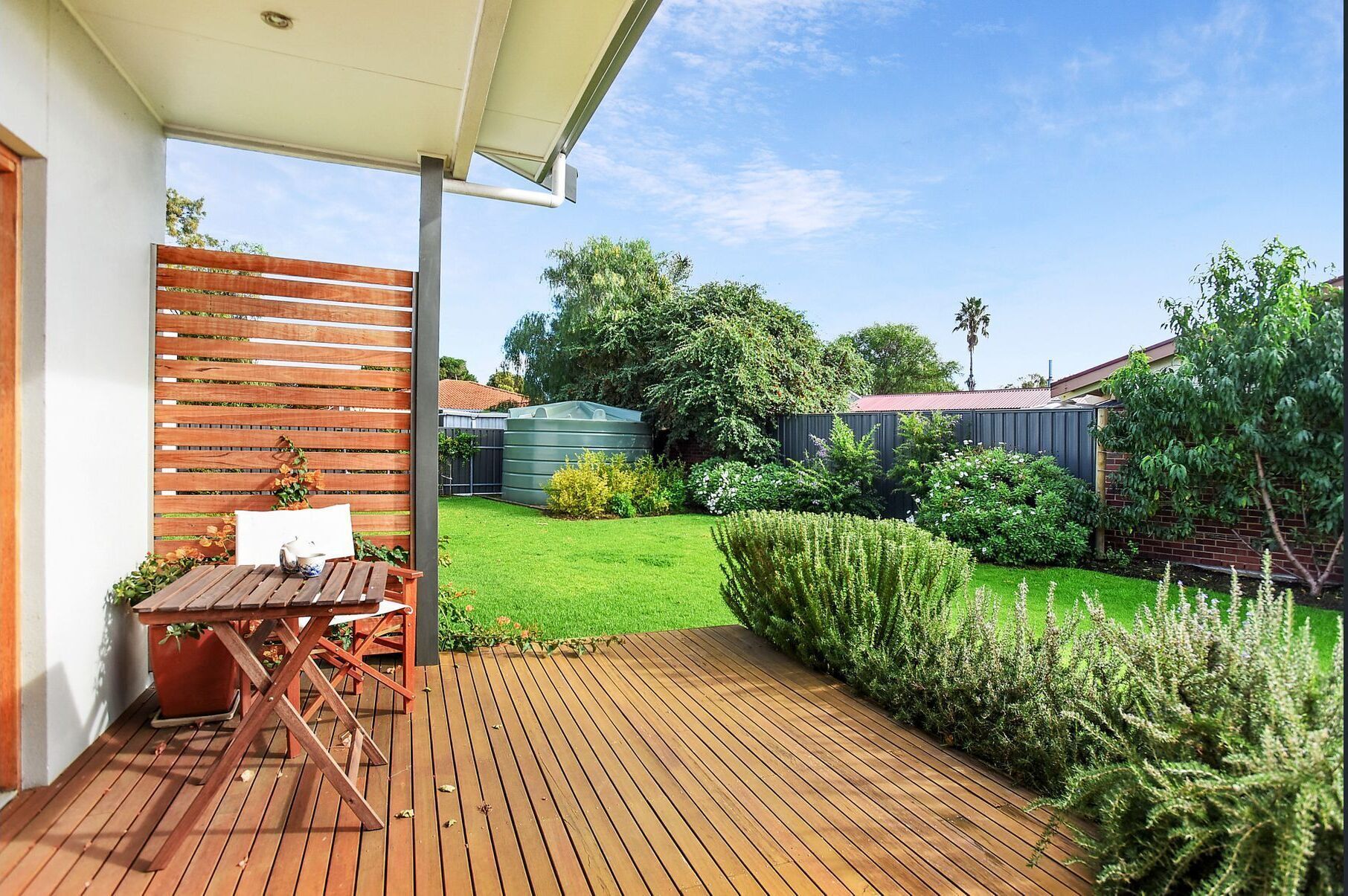A wooden deck with a table and chairs, surrounded by a well-maintained garden with various plants and a lawn.