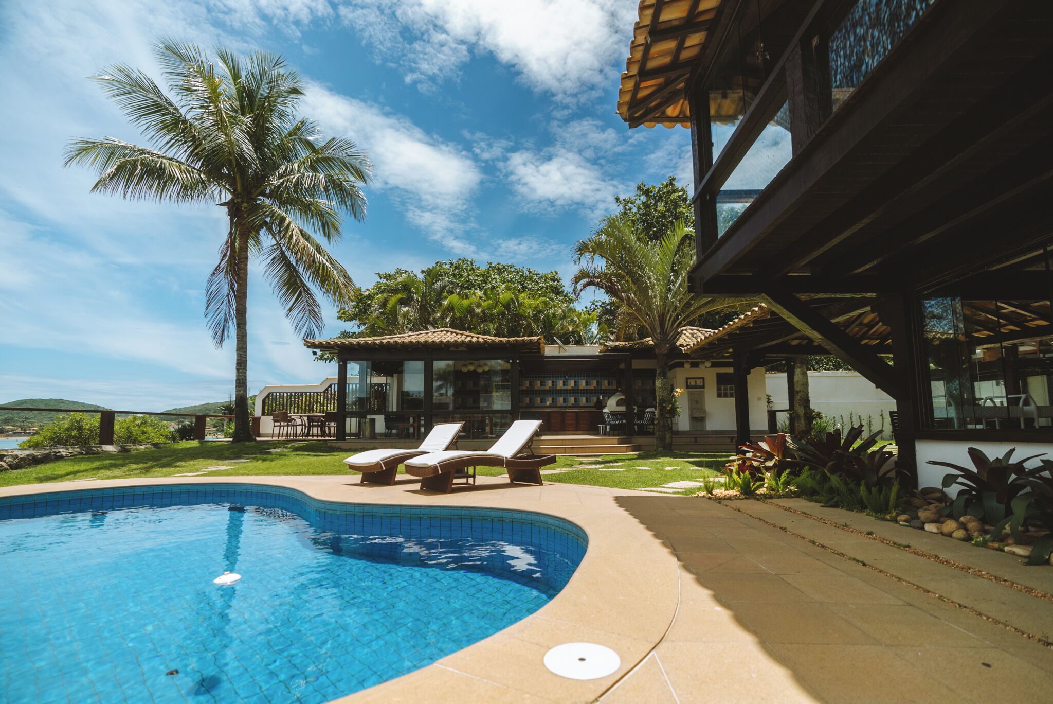 En swimmingpool med liggestole, en overdækket terrasse og udsigt til et hus med terrasse og havemøbler.