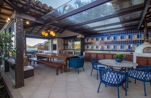 Un patio al aire libre con mesa y sillas de madera, una zona cerrada con vidrio y una cocina con azulejos azules y amarillos.