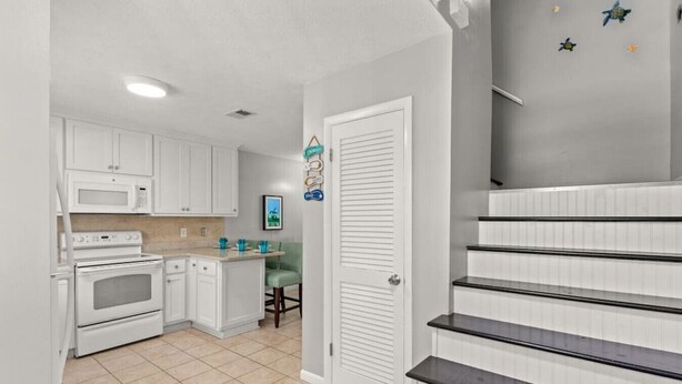 A kitchen with white cabinets, a microwave, and an oven. There is a small island with a chair and a picture on the wall.