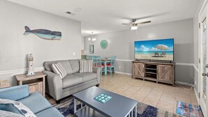 A living room with a grey sofa, a coffee table, a TV on a wooden stand, a dining area with a table and chairs, and a ceiling fan.