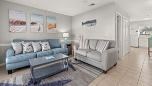 A living room with two sofas, a coffee table, and a rug. There are wall art pieces and a kitchen in the background.