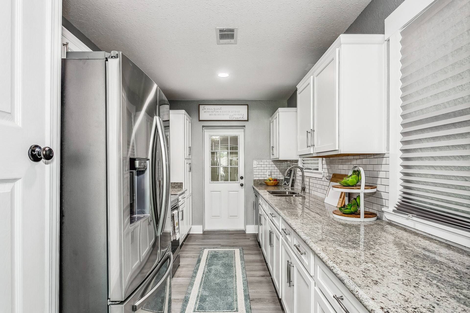 A kitchen with white cabinets, a stainless steel refrigerator, a granite countertop, and a window with blinds.