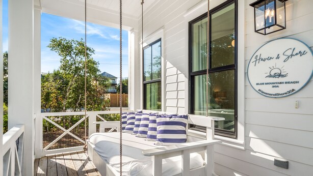 Un portico bianco con un'altalena, un letto con biancheria bianca e cuscini a righe blu, e un cartello con la scritta "Haven & Shore Blue Mountain Beach 30A".