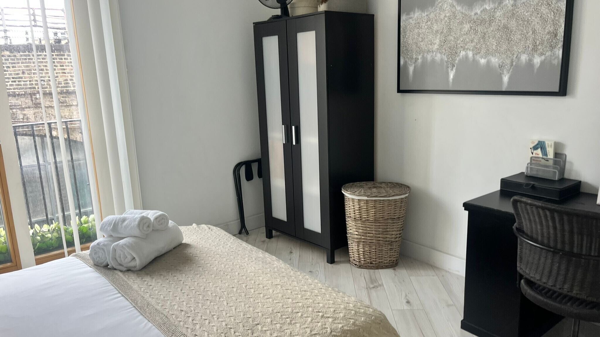 A bedroom with a bed, a wardrobe, a desk, a chair, a waste basket, and a window with a view of greenery.