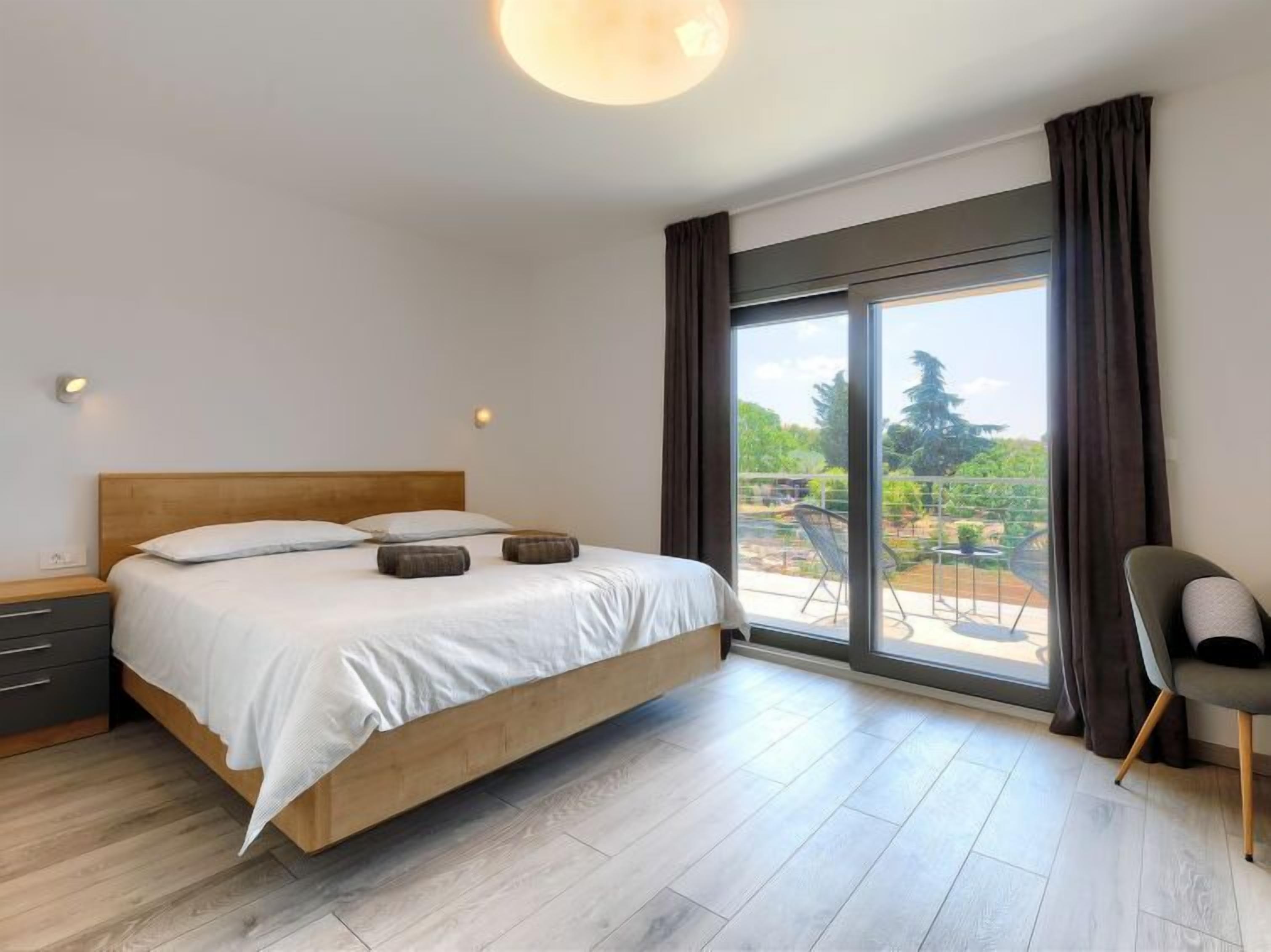 Camera da letto con un letto di legno, un comodino, una sedia e un'ampia finestra che si affaccia su una vista verde.