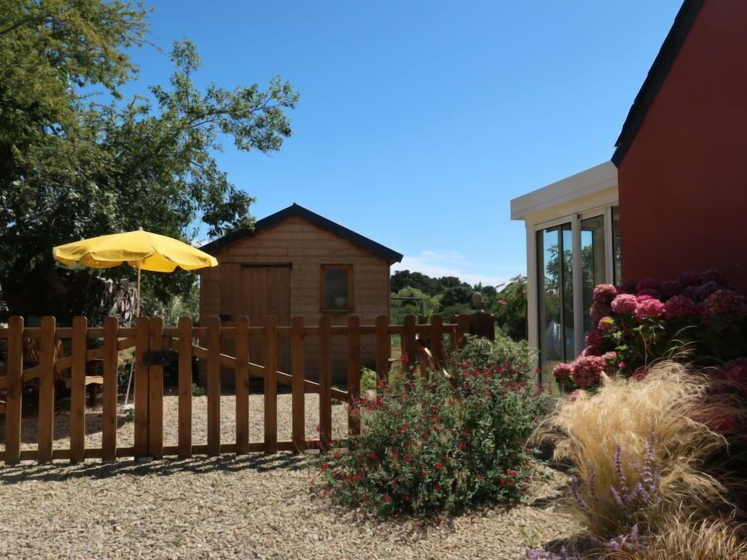 A wooden cabin with a fenced yard, a yellow umbrella, and various plants.