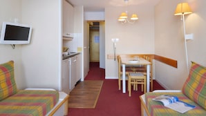 Une chambre d’hôtel compacte comprenant un coin cuisine, un espace repas avec une table et des chaises, un téléviseur fixé au mur et un canapé agrémenté d’un coussin à carreaux.