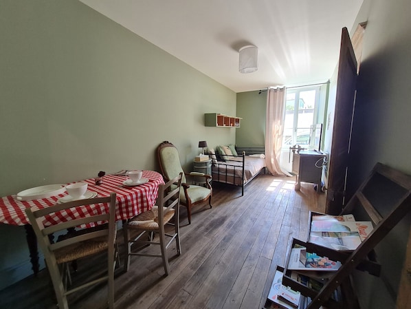 A room with a dining table set for two, a bed with a nightstand, a chair, and a small table with books.