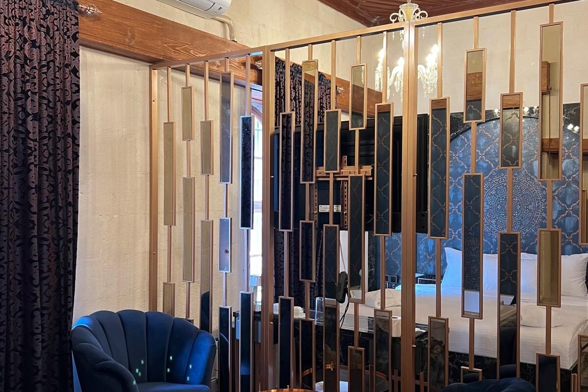 Ein modernes Hotelzimmer mit Holzbalkendecke, einer Chandelierleuchtre, einem Bett mit Kopfteil, zwei blauen Sesseln, einem runden Couchtisch und einem dekorativen Paravan mit Spiegeln.
