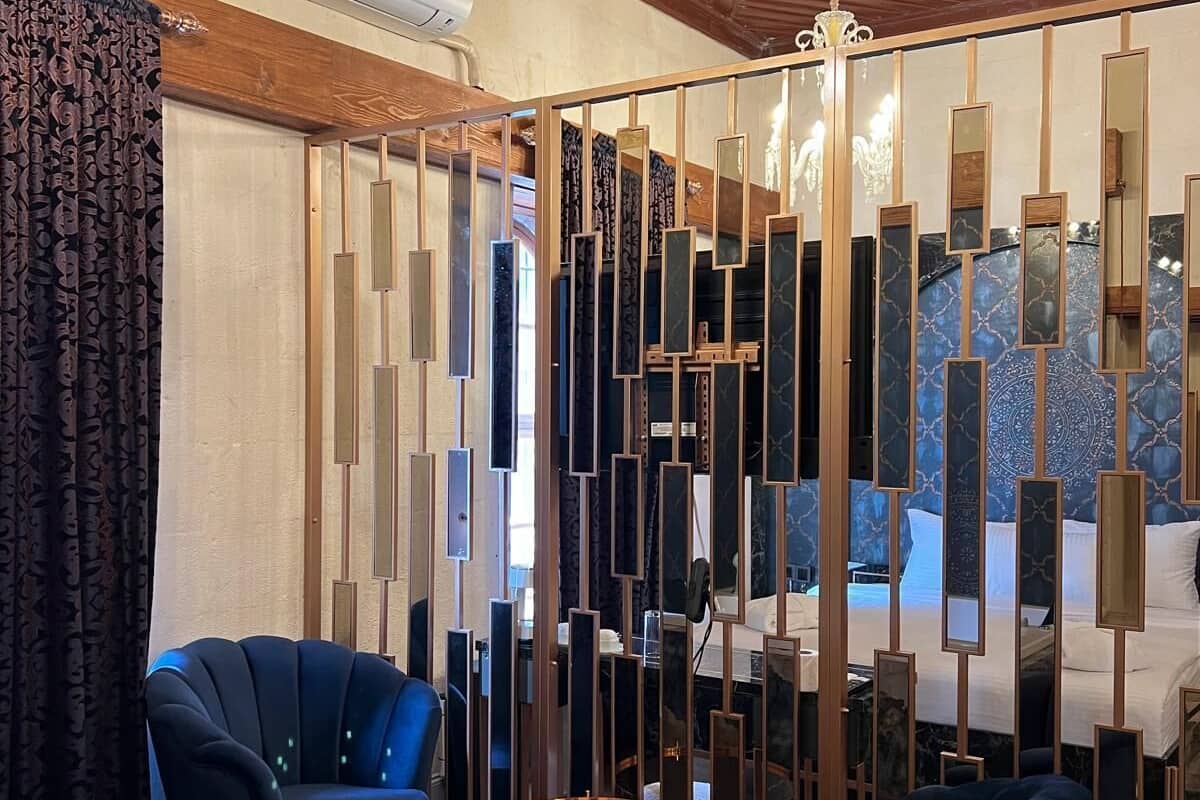 Ein modernes Hotelzimmer mit Holzbalkendecke, einer Chandelierleuchtre, einem Bett mit Kopfteil, zwei blauen Sesseln, einem runden Couchtisch und einem dekorativen Paravan mit Spiegeln.