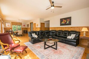 A living room with a black leather sectional sofa, a glass coffee table, and a red leather armchair. There is a wooden floor, a ceiling fan, and a painting on the wall.