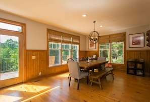 A spacious dining area with a wooden table and chairs, large windows with blinds, and a chandelier.