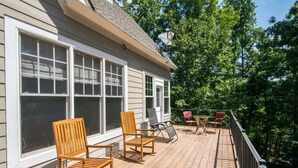 A wooden deck with outdoor furniture, including chairs and a table, adjacent to a house with large windows. - Lake Adger Home with Private Dock & Kayaks (Mill Spring)