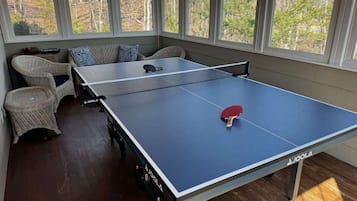 Mesa de ping pong en una habitación con ventanas amplias, mobiliario de mimbre y vistas a árboles.