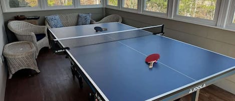 Mesa de ping pong en una habitación con ventanas amplias, mobiliario de mimbre y vistas a árboles.