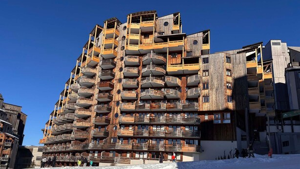 Un edificio de varios pisos con balcones, una pista de esquí en primer plano y un cielo azul despejado.