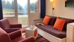 Una habitación moderna con sofá, dos sillones, una mesita con tazas, televisión y ventana con vistas a árboles y una valla.