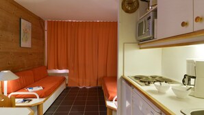 A compact kitchen with a stove, microwave, and sink. There is a wooden table with a lamp and a sofa with orange cushions.