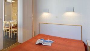 A hotel room with a bed, a table with a magazine, a chair, a wooden door, and a mirror.
