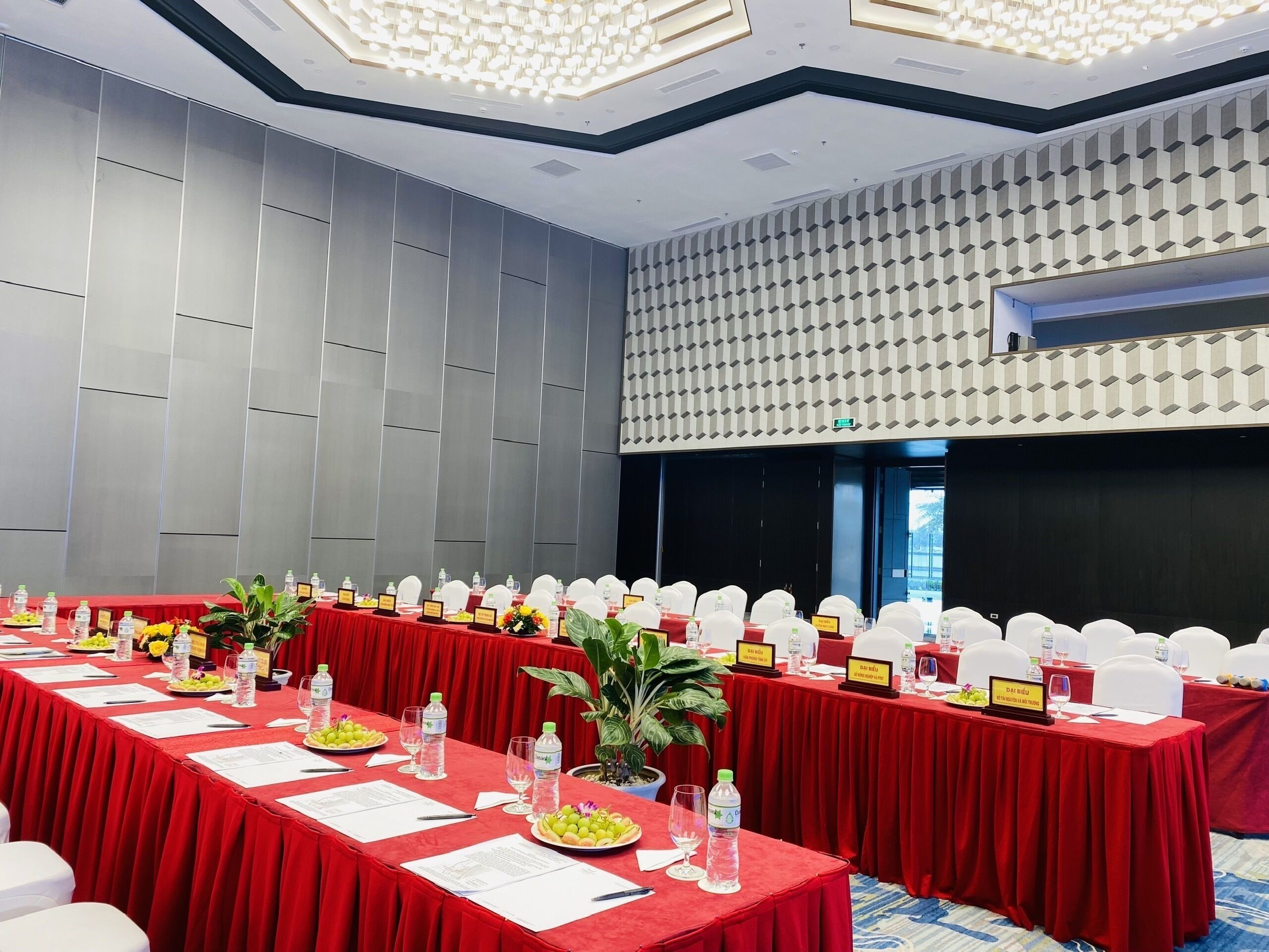 A conference room with red tables, white chairs, nameplates, water bottles, and potted plants.