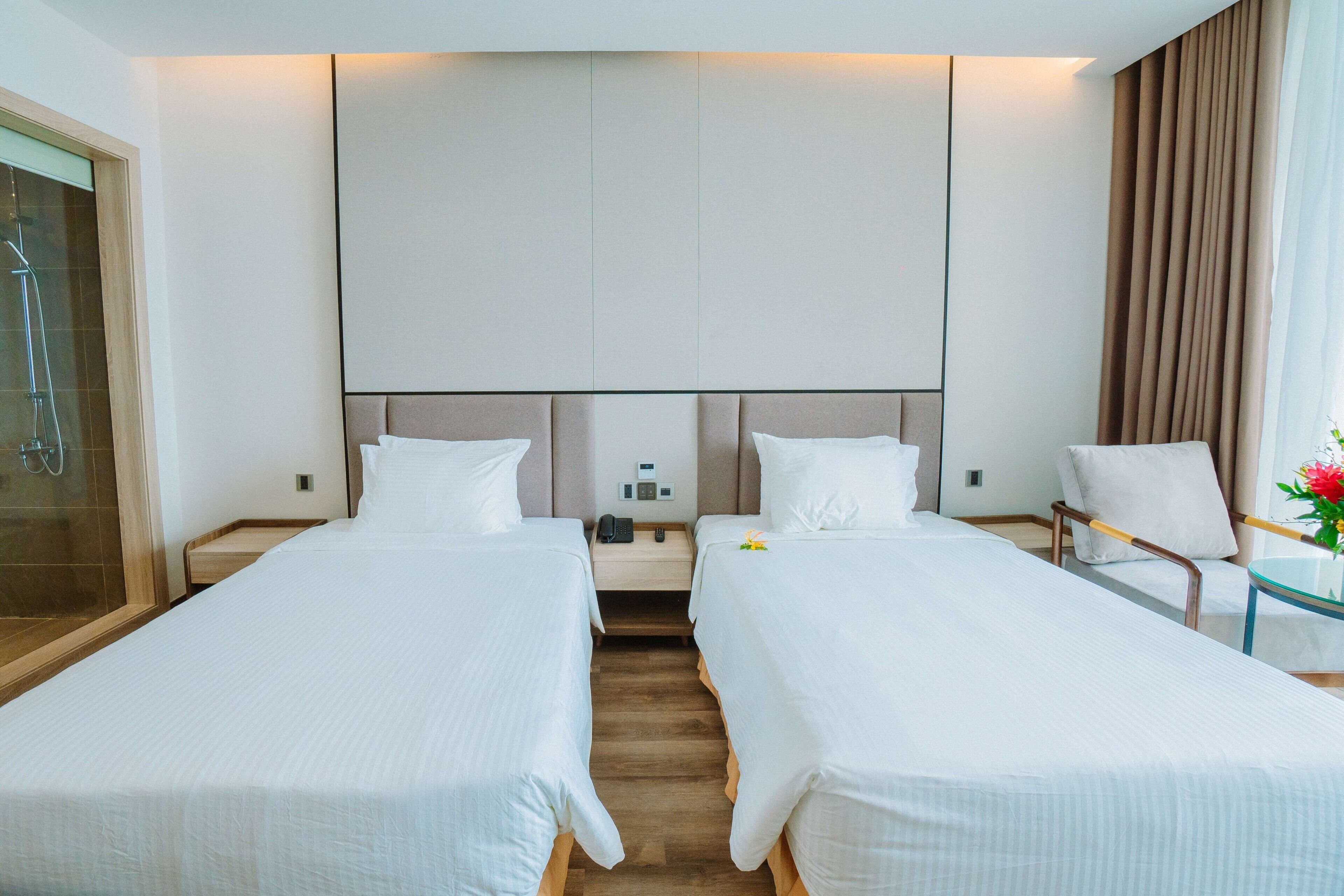 Ein Hotelzimmer mit zwei Einzelbetten, einem Nachttisch mit Telefon und Lampe, einem Sessel und einem Glastisch mit Blumenvase.