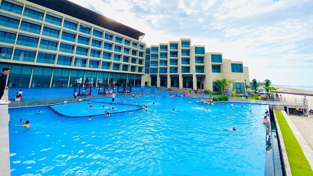 A large outdoor swimming pool area with multiple pools and a modern building with large windows in the background. - Muong Thanh Luxury Xuan Thanh Hotel (Tien Dien)