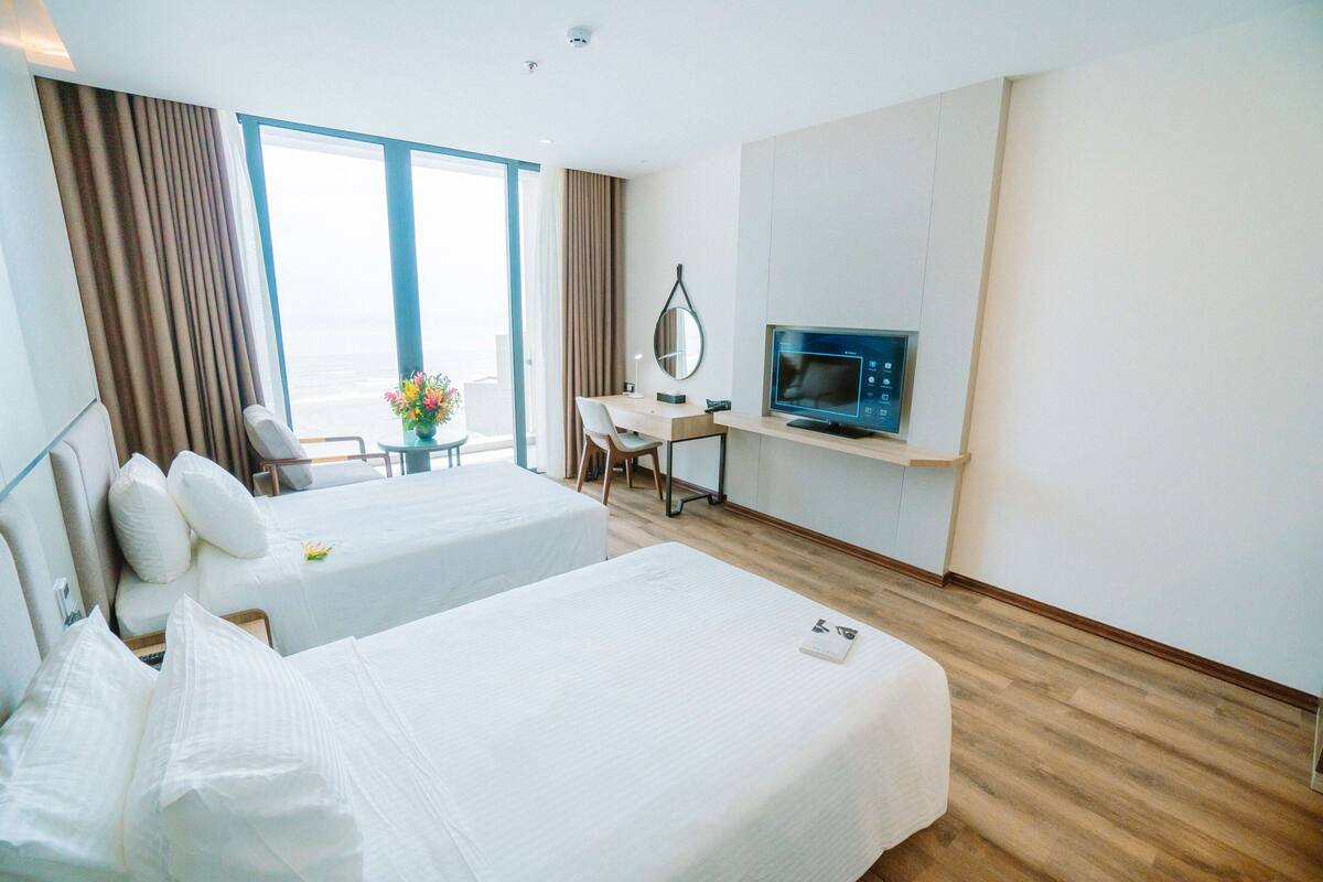 Ett hotellrum med två sängar, ett skrivbord med en dator, en tv och ett stort fönster med gardiner.