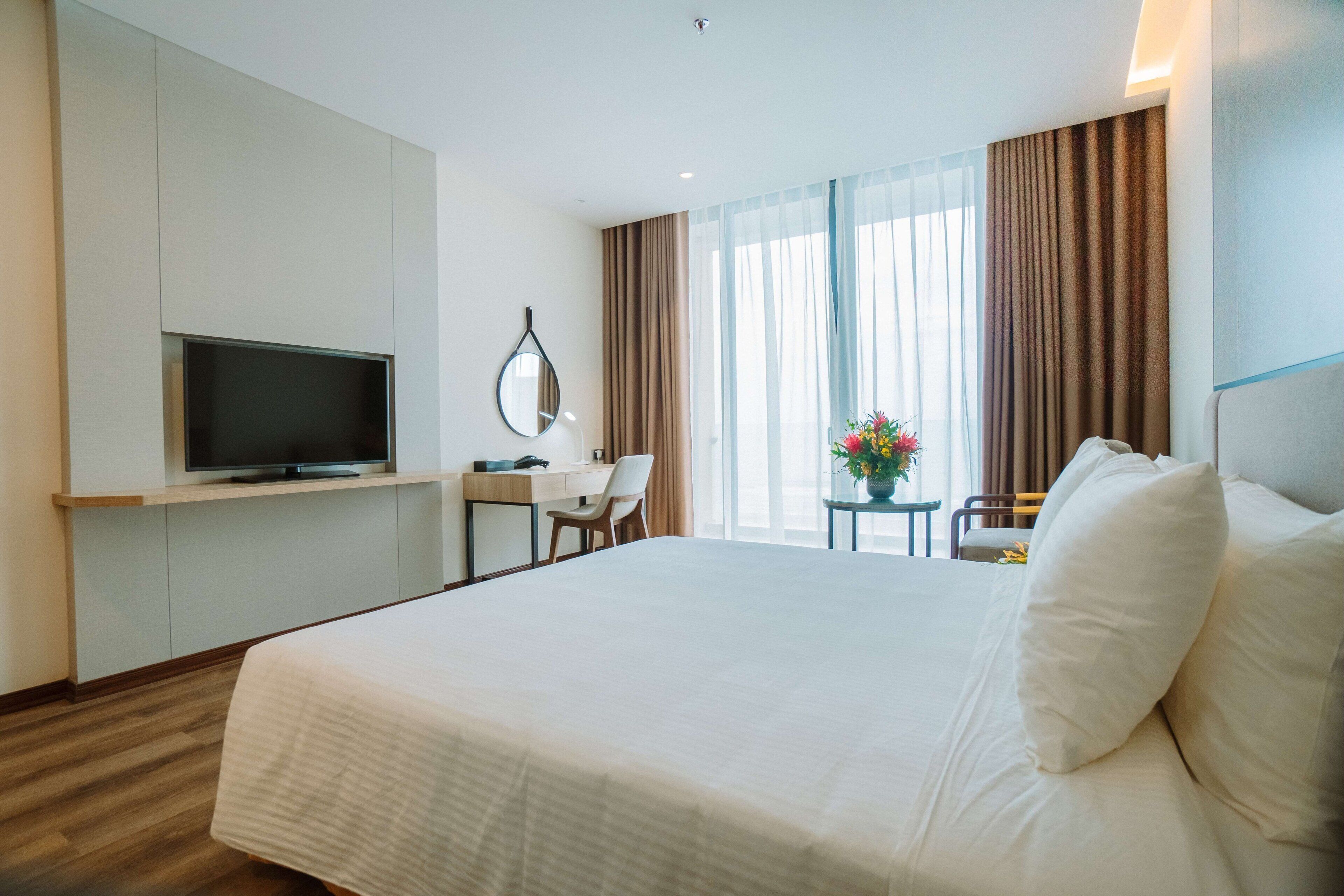 Quarto de hotel com uma cama grande, uma escrivaninha com cadeira, televisão e uma janela com cortinas.