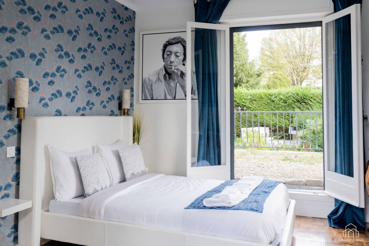 A bedroom with a bed, a window with curtains, a wall with a patterned wallpaper, and a framed picture on the wall.