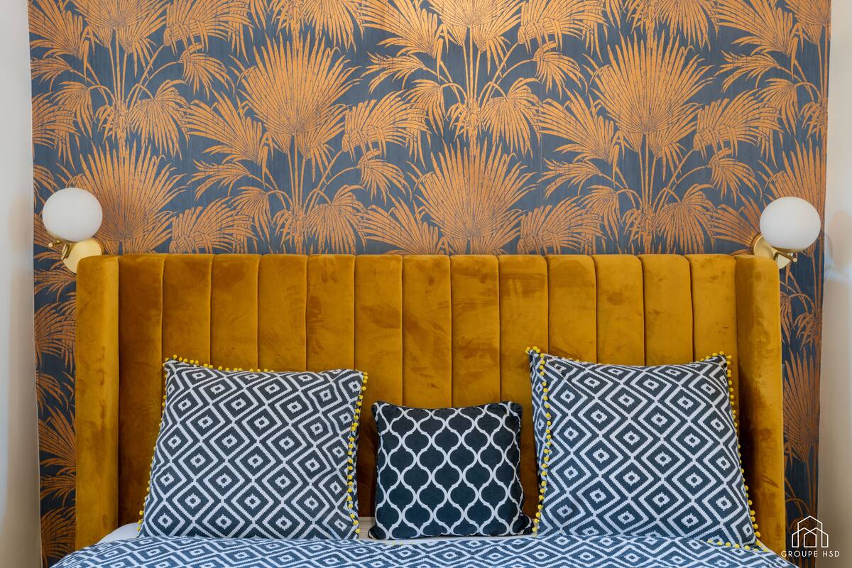 A bed with a yellow headboard, patterned pillows, and a floral wallpaper background.
