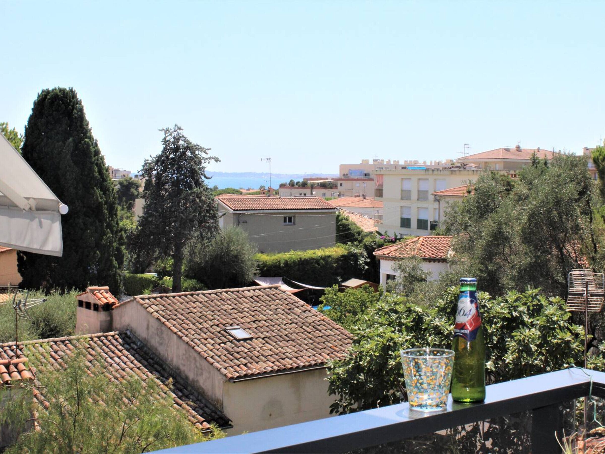 Un quartier résidentiel aux toits recouverts de tuiles en terre cuite, offrant une vue dégagée sur la mer, et doté d’un balcon avec une bouteille et un verre.