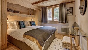 Un dormitorio acogedor con techo de madera, una cama grande con una manta de diseño en espiga, una ventana con cortinas y una mesita de noche con lámpara.