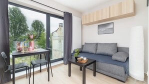 A modern living room with a grey sofa, a small table with a plant, and a window overlooking a garden. - Stylish Studio in Krakow | Air Conditioning | Close to the Station (Kraków)