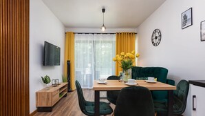 Een moderne eethoek met een houten tafel, groene stoelen, een groene bank, een aan de muur bevestigde televisie en een raam met gele gordijnen.