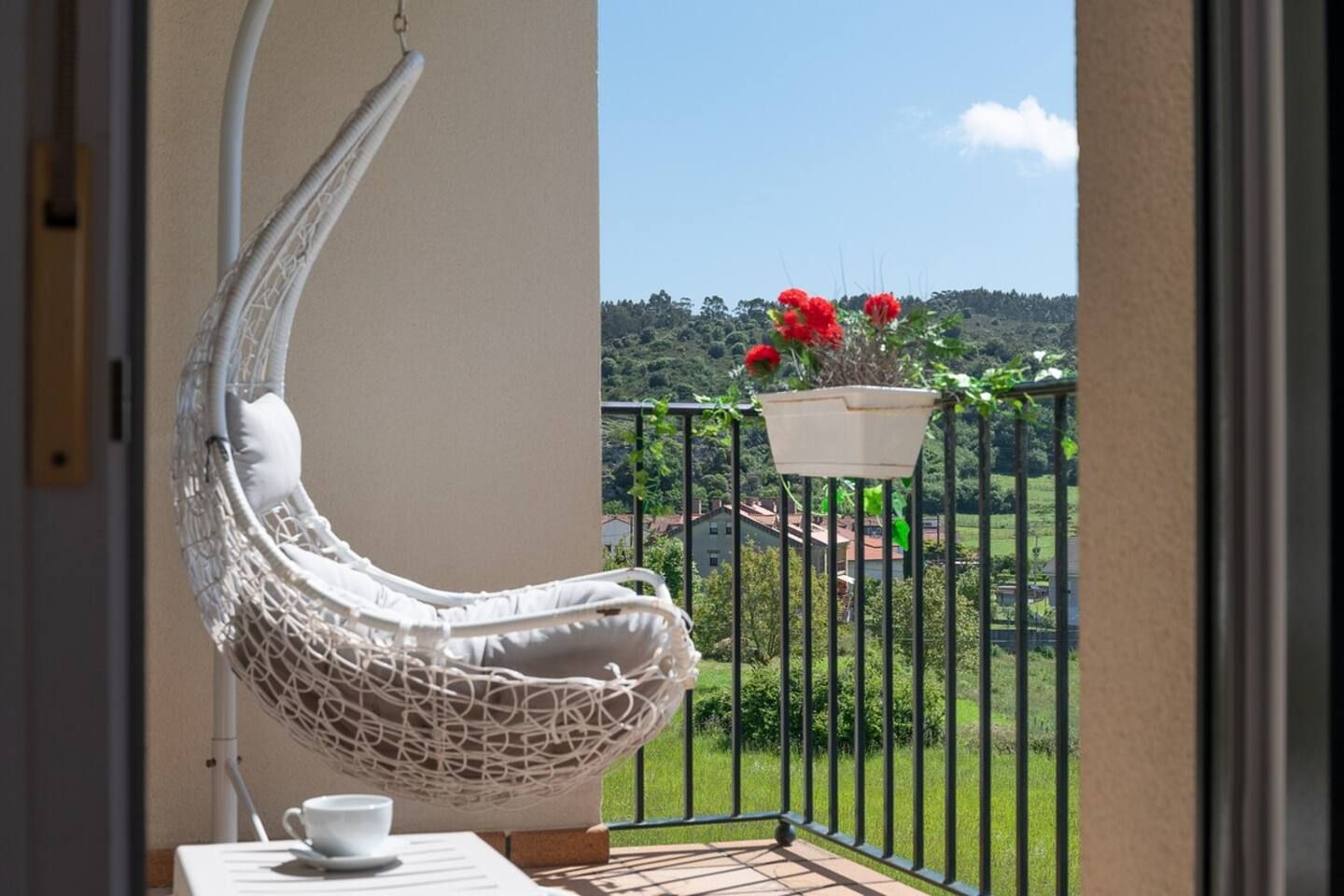 Una silla colgante blanca en un balcón con vistas a un paisaje verde y una maceta roja.