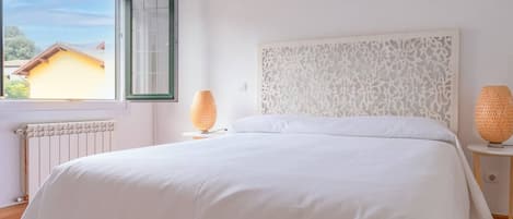 Quarto com cama grande, janela com vista para um edifício, radiador e duas luminárias sobre mesas de cabeceira.