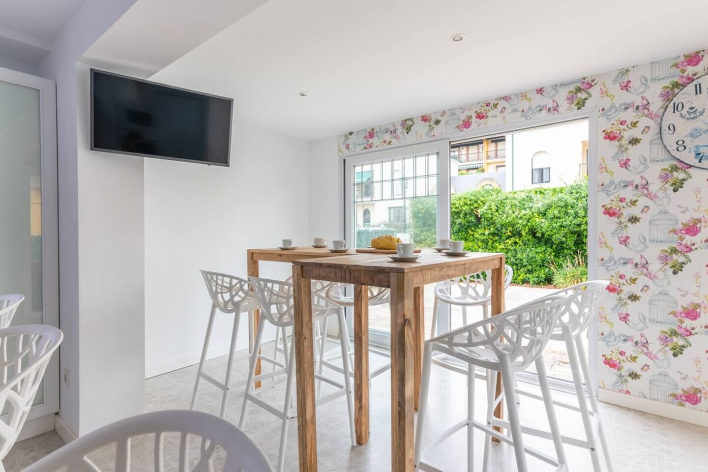 Un coin repas avec une table en bois et des chaises blanches, un téléviseur à écran plat fixé au mur, et une fenêtre donnant sur un paysage verdoyant et un bâtiment.