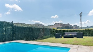 A swimming pool with a clear blue water surface, surrounded by a paved deck. In the background, there is a grassy area with a hedge and a view of a hill with a power line tower.
