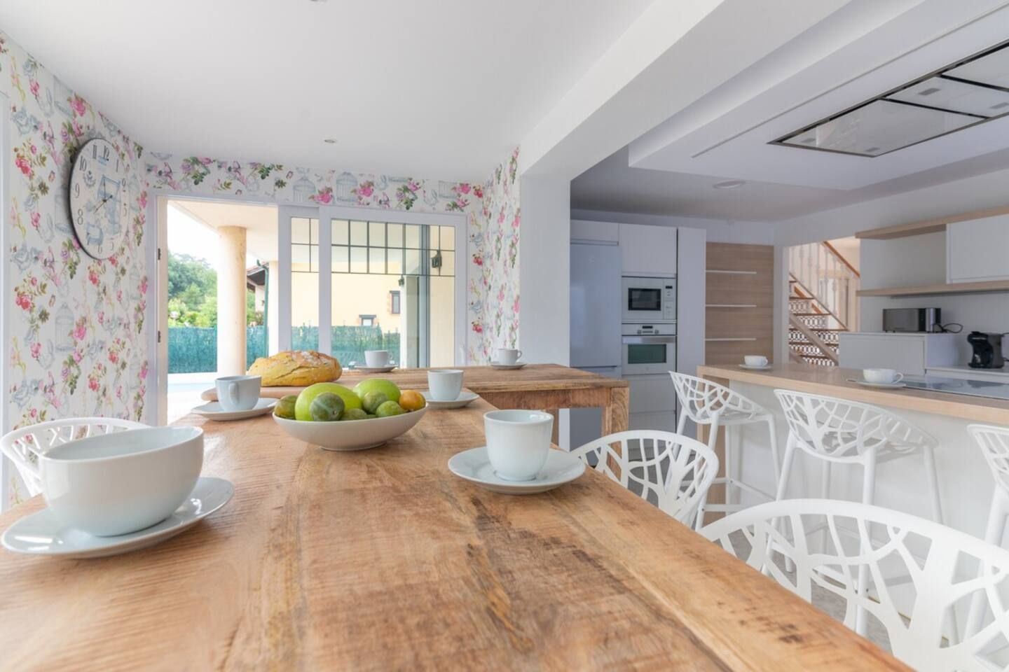A dining area with a wooden table set for breakfast, white chairs, and a bowl of fruit.