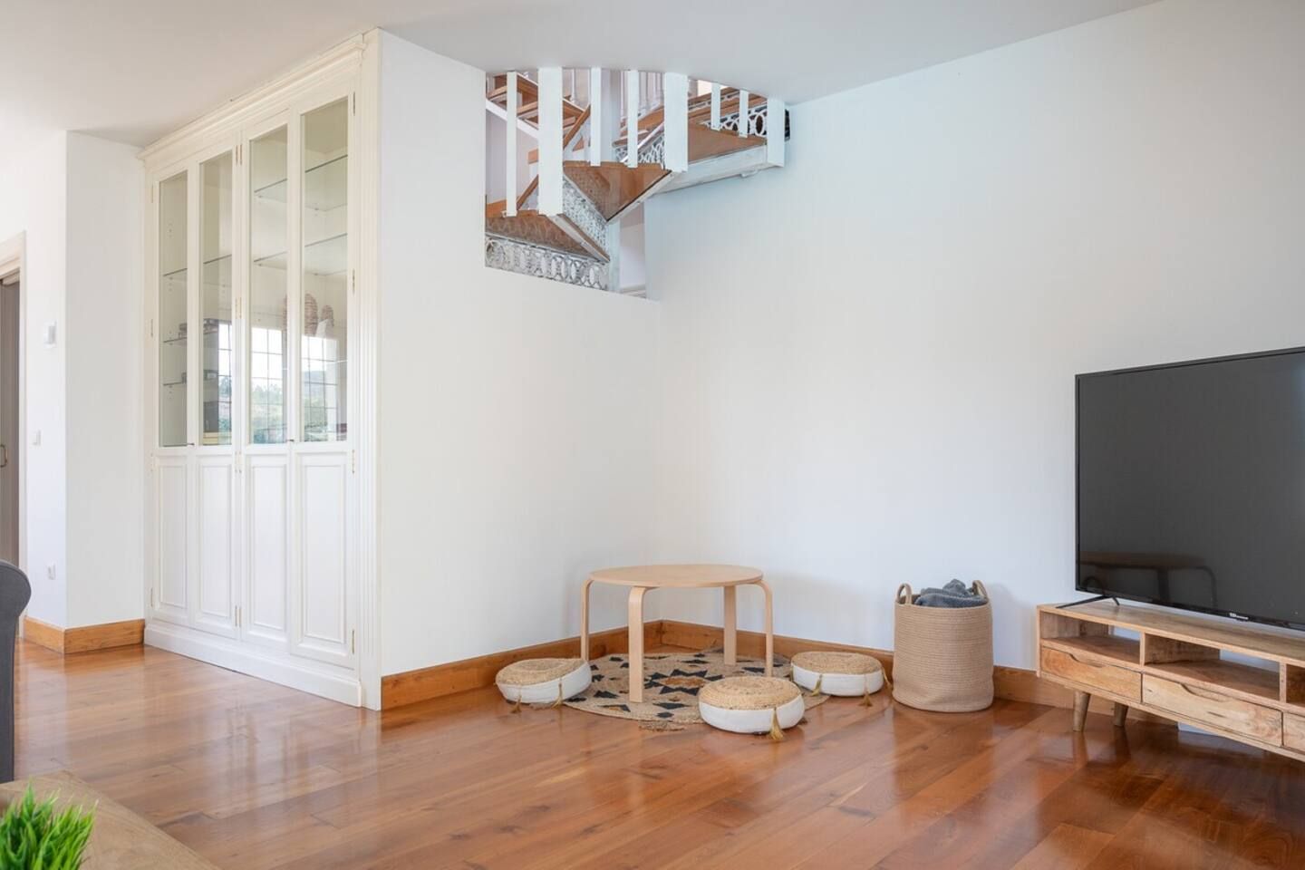 Un salon moderne avec un plancher en bois, un téléviseur, un meuble blanc aux portes vitrées, une petite table ronde et un escalier.